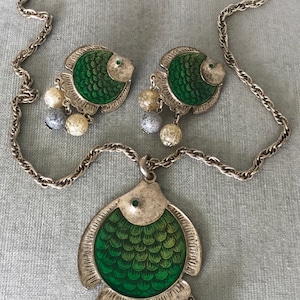 Puede incluir: Un collar de cadena de plata con un colgante de pez verde y plateado. El colgante tiene un acabado de esmalte verde y está decorado con pequeñas escamas plateadas. El collar también incluye un par de pendientes de pez verde y plateado a juego.