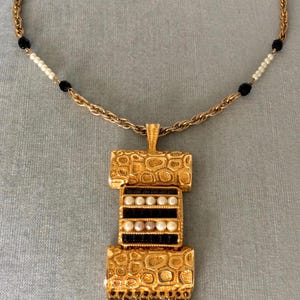 Puede incluir: Un collar de cadena dorada con un gran colgante que presenta un diseño dorado texturizado, cuentas negras y blancas y un borla de cuentas doradas y negras.