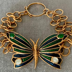 May include: Gold-toned butterfly bracelet with green, blue, and white enamel wings. The butterfly has a detailed design with a chain link band. The bracelet features a clasp for secure wear and is a statement piece.