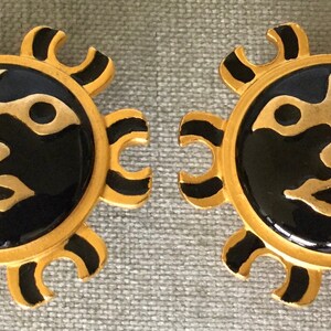 May include: A pair of gold-toned sun-shaped earrings with black enamel centers. The sun design features a face with gold details and black accents. The earrings are likely clip-ons or post-style.