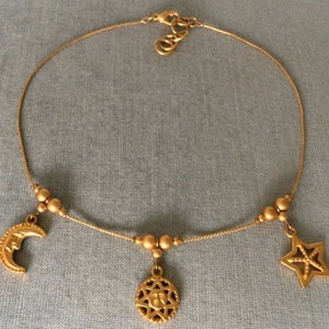 May include: A gold chain necklace with three charms: a crescent moon, a star, and a sun with a face. The charms are connected to the chain with small gold beads.