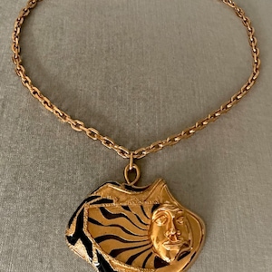 May include: Gold-tone necklace featuring a pendant with a stylized face and abstract design. The pendant has black and green accents. The chain is a linked design. The necklace is displayed on a neutral background.