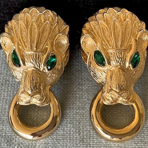 Op de afbeelding: Een paar goudkleurige leeuwenkop oorbellen met groene edelsteen ogen en een ring detail. De oorbellen hebben een getextureerde afwerking en zijn ontworpen om als clip-ons of oorstekers te worden gedragen.