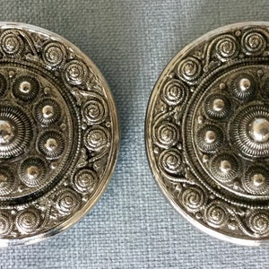 May include: A pair of silver-toned metal buttons with a Celtic design. The buttons feature a central circle surrounded by smaller circles and a decorative border of swirling patterns.