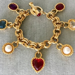 May include: A gold-tone chain link bracelet featuring a toggle clasp and six dangling charms. The charms include two white pearls, two red heart-shaped stones, one blue heart-shaped stone, and one oval purple stone, all set in twisted gold-tone metal frames.