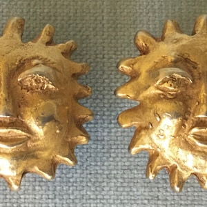 May include: A pair of gold-colored sun-shaped earrings. Each earring features a detailed face with closed eyes, a small nose, and a closed mouth, surrounded by spiky rays. The earrings are likely made of metal.