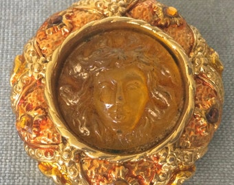 May include: A gold-toned brooch with a round, raised, amber-colored cameo of a woman's face. The cameo is surrounded by a gold-toned frame with a textured design. The brooch is decorated with small, clear and amber-colored rhinestones.