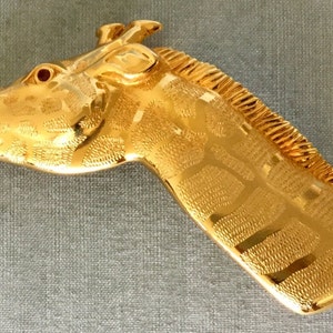Könnte beinhalten: Eine goldfarbene Giraffenkopf-Brosche mit detaillierter, strukturierter Oberfläche. Die Brosche hat ein rotes Strassstein-Auge.
