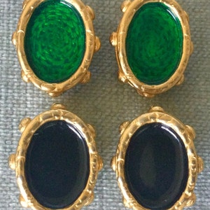 May include: Four vintage gold-toned clip-on earrings. Two earrings have oval, emerald green centers, and two have black centers. Each earring has a decorative gold border with small, raised details. The earrings are displayed on a light blue-green surface.