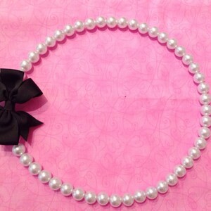 Puede incluir: Un collar de perlas blancas con un lazo negro sobre un fondo rosa.