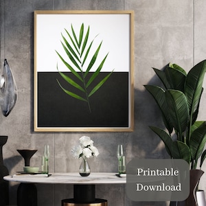 May include: A printable art print featuring a single green palm leaf on a black and white background. The print is framed in a light wood frame and is hanging on a gray wall.