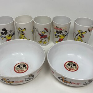 May include: Set of five white plastic cups and two white plastic bowls featuring Mickey Mouse, Donald Duck, Pluto, and Goofy characters. The cups and bowls have a red and yellow Mickey Mouse Club Cereal Bowl label.