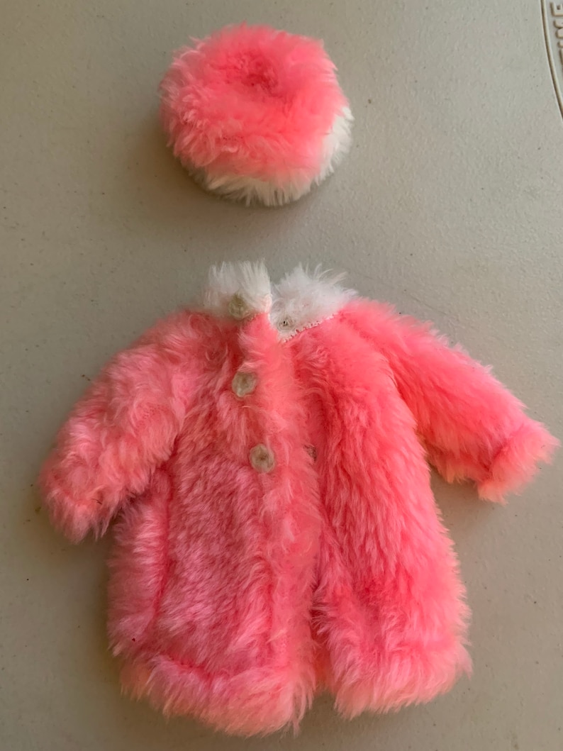 May include: A pink faux fur coat with white trim and buttons. The coat is paired with a matching pink faux fur hat with a white trim.