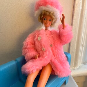 May include: A pink fur coat with a white fur trim and a matching pink hat with a white fur trim. The doll is sitting on a blue couch.