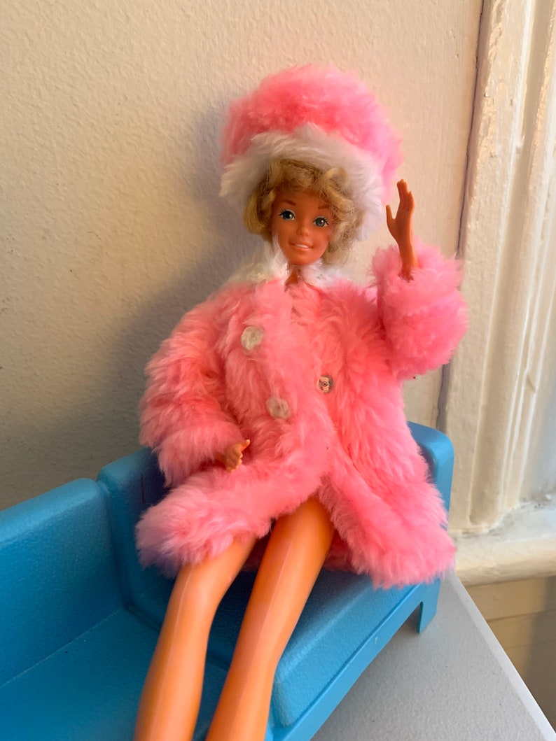 May include: A pink fur coat with a white fur trim and a matching pink hat with a white fur trim. The doll is sitting on a blue couch.