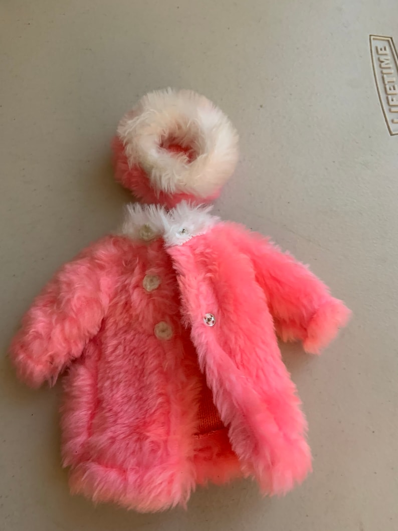 May include: A pink faux fur coat with white trim and three white buttons. The coat has a matching white faux fur hat.