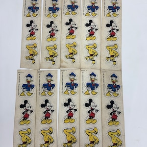 May include: A sheet of vintage Disney character stickers featuring Mickey Mouse, Donald Duck, and Pluto. The stickers are printed on white paper with a light brown background. The characters are depicted in various poses, including Mickey Mouse standing with his hands on his hips, Donald Duck wearing a sailor hat, and Pluto standing on his hind legs.