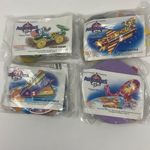 May include: Four sealed McDonald's Happy Meal toys featuring astronaut-themed designs. The toys include a Lunar Rover, Space Shuttle, Command Module, and Satellite Dish. Each toy is packaged in a clear plastic bag with the McDonald's Astronauts Happy Meal logo.