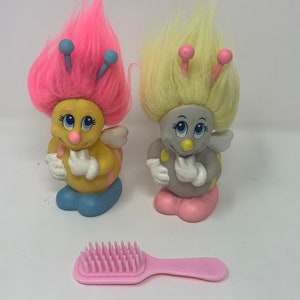 May include: Two small plush toys shaped like bees with pink and yellow hair, and a pink plastic comb. The bee toys have blue wings and pink and yellow accents.