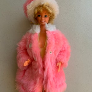 May include: A vintage Barbie doll wearing a pink faux fur coat and a white and pink hat. The doll has blonde hair and blue eyes.