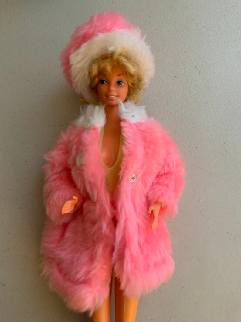 May include: A vintage Barbie doll wearing a pink faux fur coat and a white and pink hat. The doll has blonde hair and blue eyes.