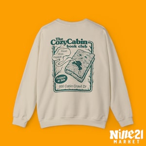 Puede incluir: Sudadera color crema con un gráfico verde en la espalda. El gráfico dice "The Cozy Cabin book club" con una ilustración de libro y el texto "Read. Rise. Repeat." y "Fridays at 3am, 666 Cabin Crawl Dr."