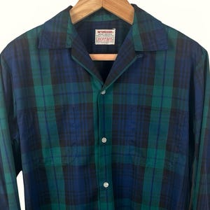 May include: A long-sleeved plaid shirt in shades of blue and green, buttoned up and hanging on a wooden hanger. The shirt has two chest pockets and a collar. The label reads "McGregor SCOTSET".