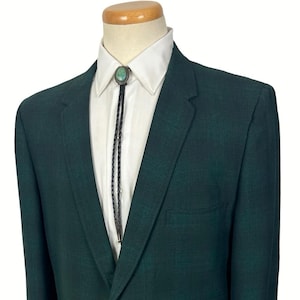 May include: A dark forest green blazer with a single button closure, notched lapels, and two flap pockets, worn over a crisp white collared shirt featuring a black braided bolo tie with a central oval turquoise stone accent.
