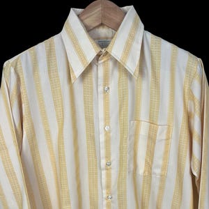 May include: A vintage cream and yellow striped button-down shirt. The long-sleeved shirt has a pointed collar, a chest pocket, and buttoned cuffs. It is displayed on a wooden hanger.