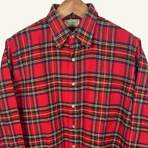 May include: Red plaid button-down shirt with long sleeves, hanging on a wooden hanger. The shirt features a classic plaid pattern in red, black, yellow, and blue. The label reads "L.L. Bean".