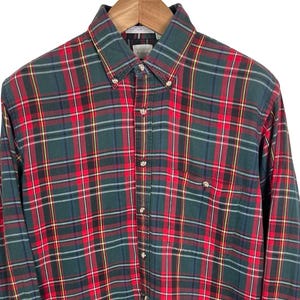 Może przedstawiać: Koszula w kratę z długim rękawem w kolorze czerwonym, zielonym, czarnym i żółtym, zapięta na guziki i wisząca na drewnianym wieszaku. Koszula ma kołnierzyk zapinany na guziki i jedną kieszeń na piersi. Wzór w kratę to klasyczny design.