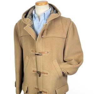 May include: A tan duffle coat featuring a hood, long sleeves, and two large front pockets. The coat is fastened with three wooden toggles and leather loops. A light blue collared shirt is visible underneath, and the hood interior has a plaid pattern.
