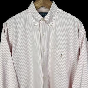 May include: A long-sleeved button-down shirt with a pink and white striped pattern. The shirt has a button-down collar, a chest pocket, and a small embroidered logo. The shirt is made of a lightweight fabric.