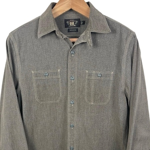 May include: A long-sleeved, button-down shirt in a muted olive green hue. The shirt features two chest pockets, a classic collar, and buttoned cuffs. The brand logo is visible on the label. The shirt is hanging on a wooden hanger.