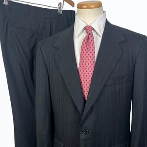 May include: A dark gray pinstripe suit with a white dress shirt and a pink patterned tie. The suit jacket has two front pockets and a two-button closure. The pants are hanging on a wooden hanger.