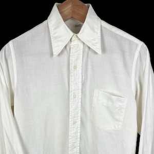 May include: Cream-coloured long-sleeved shirt with a pointed collar, button-down front, and a chest pocket. The shirt is hanging on a wooden hanger. The fabric appears to be a lightweight material.
