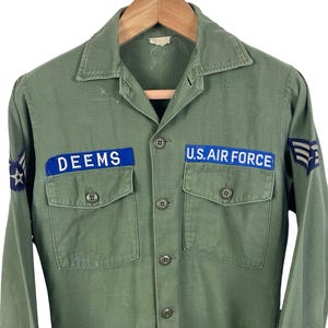 May include: An olive green long-sleeve button-up shirt featuring two chest pockets, a collar, and blue patches with white text reading DEEMS and U.S. AIR FORCE. The sleeves include embroidered insignia patches on each shoulder area.