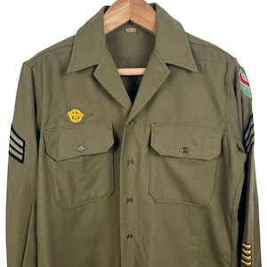 May include: An olive green long-sleeve military-style shirt featuring a pointed collar, a button-down front, and two chest pockets. The shirt includes embroidered patches on the sleeves and a small yellow emblem on the right chest area.