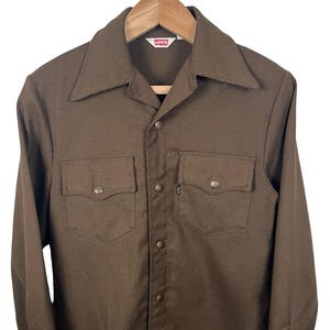 May include: A brown button-down shirt with two chest pockets and a collar. The shirt has long sleeves and is hanging on a wooden hanger. The Levi's label is visible at the neck.