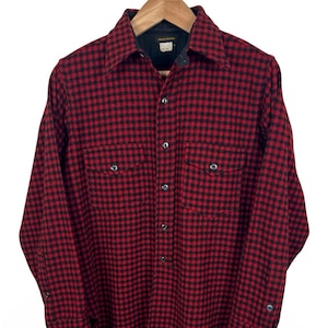 Pode incluir: Uma camisa de flanela xadrez vermelha e preta com mangas compridas, dois bolsos no peito e uma frente abotoada. A camisa está pendurada em um cabide de madeira.