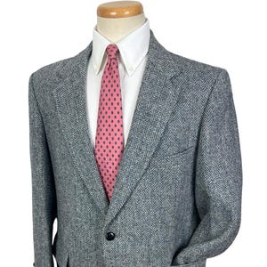Puede incluir: Un blazer gris con estampado de espiga, camisa blanca y corbata rosa con lunares azul marino. El blazer tiene dos bolsillos delanteros y un cierre de un solo botón. El blazer se exhibe en un maniquí.