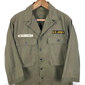 May include: An olive green long-sleeve button-up jacket featuring a collar, two front chest pockets with buttons, and a central button closure. A black patch with yellow text reads U.S. ARMY, and a white name tape reads KIRCHOFF.