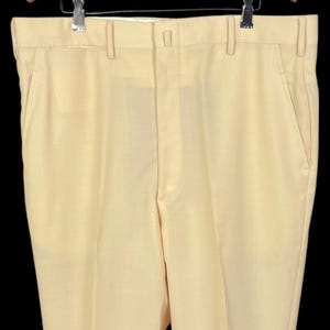 Puede incluir: Pantalones amarillo pálido con un diseño clásico de pierna recta. Los pantalones tienen trabillas, bolsillos delanteros y cierre de botón. Los pantalones cuelgan de una percha de madera sobre un fondo negro.