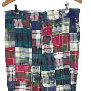 May include: Patchwork plaid pants in a variety of colors, including red, green, blue, and white. The pants are made of fabric squares sewn together in a patchwork design. The pants are hanging on a wooden hanger.