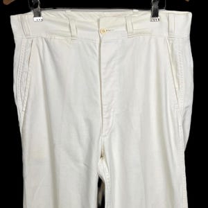 Puede incluir: Pantalones color crema con diseño de pierna recta. Los pantalones tienen cierre de botón y cremallera, trabillas para el cinturón y bolsillos delanteros y traseros. Colgados de una percha de madera.