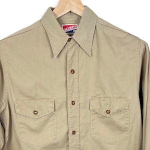 May include: A long-sleeved, light brown button-down shirt with two chest pockets and a red and white "Western Wear" label on the collar.