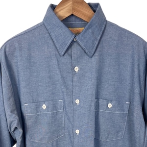 May include: A light blue long-sleeve shirt with two chest pockets and white buttons. The shirt is made of a textured fabric and hangs on a wooden hanger.