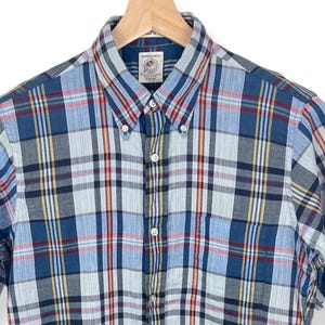 May include: A blue, red and white plaid short-sleeved button-down shirt with a small pocket on the left chest. The shirt has a button-down collar and a button closure.