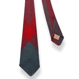 Può includere: Una cravatta fantasia rossa, grigia e grigio scuro. La cravatta presenta un motivo a rombi rossi e grigi, con una sezione grigio scuro nella parte inferiore. Una piccola etichetta è visibile vicino all'estremità della cravatta.