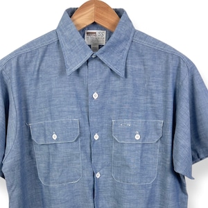 Puede incluir: Camisa azul claro de manga corta con dos bolsillos en el pecho y botones blancos. La camisa está colgada de una percha de madera. Tiene un cuello clásico y un corte relajado. La tela parece ser una mezcla ligera de algodón.